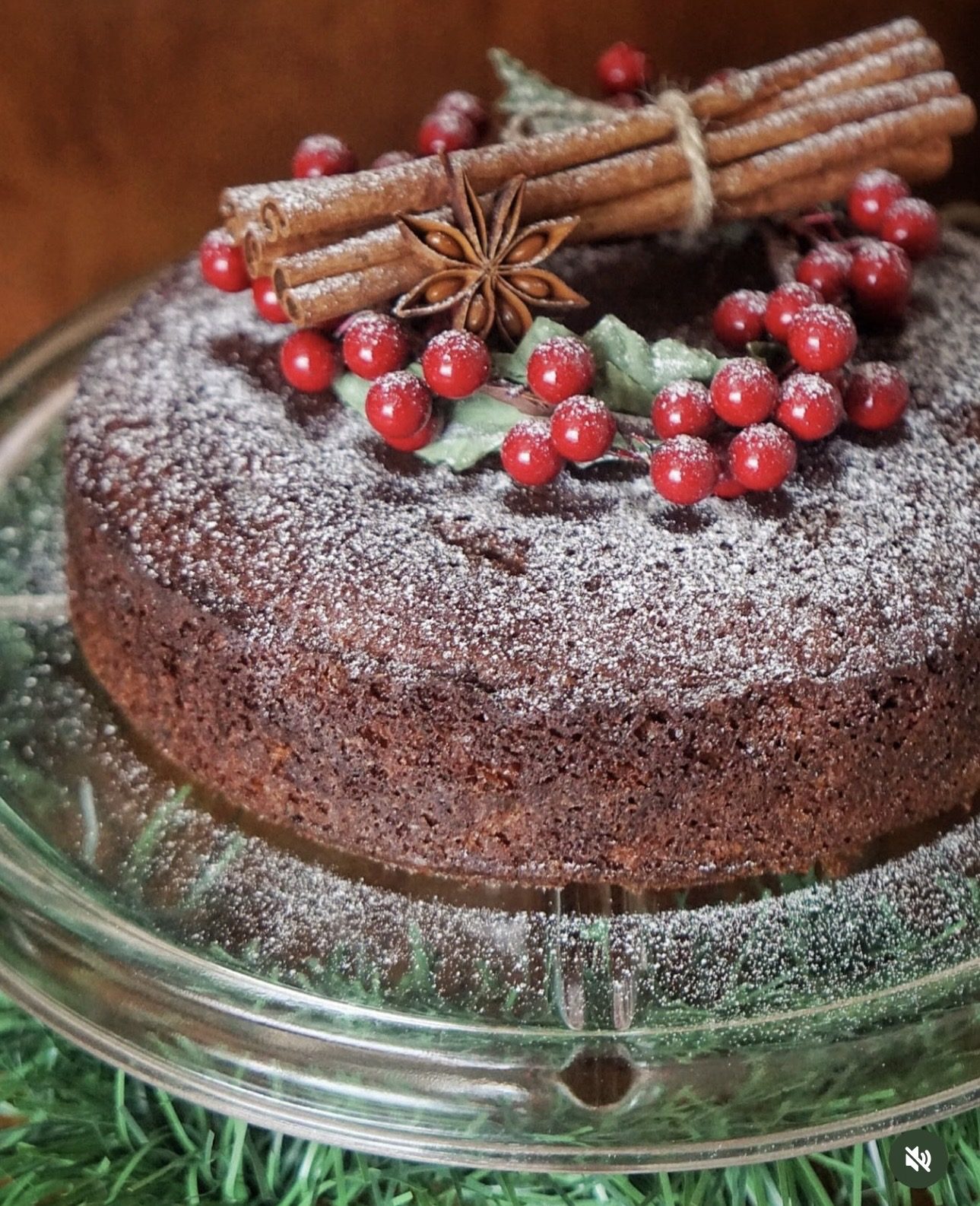 Christmas fruit cake dusted with icing sugar and decorated with cinnamon sticks, star anise and red berries on a glass cake stand.