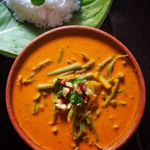 Colocasia and amaranth stalk curry in a clay bowl, topped with garlic, mustard seeds and curry leaves, with white rice behind it.
