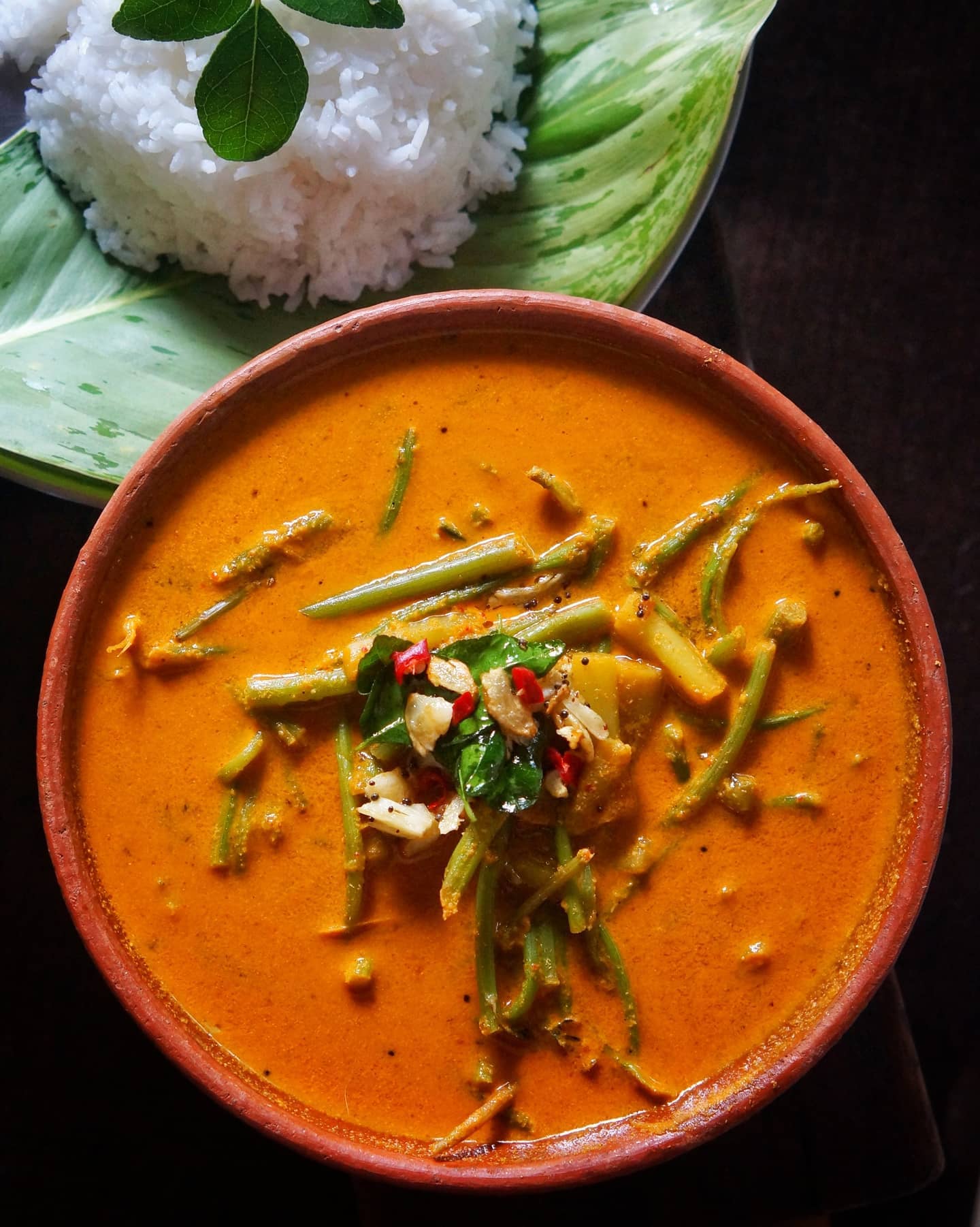 Colocasia and amaranth stalk curry in a clay bowl, topped with garlic, mustard seeds and curry leaves, with white rice behind it.