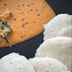 Orange-red Kempu Chutney with mustard seeds and curry leaves beside white idlis on a dark plate