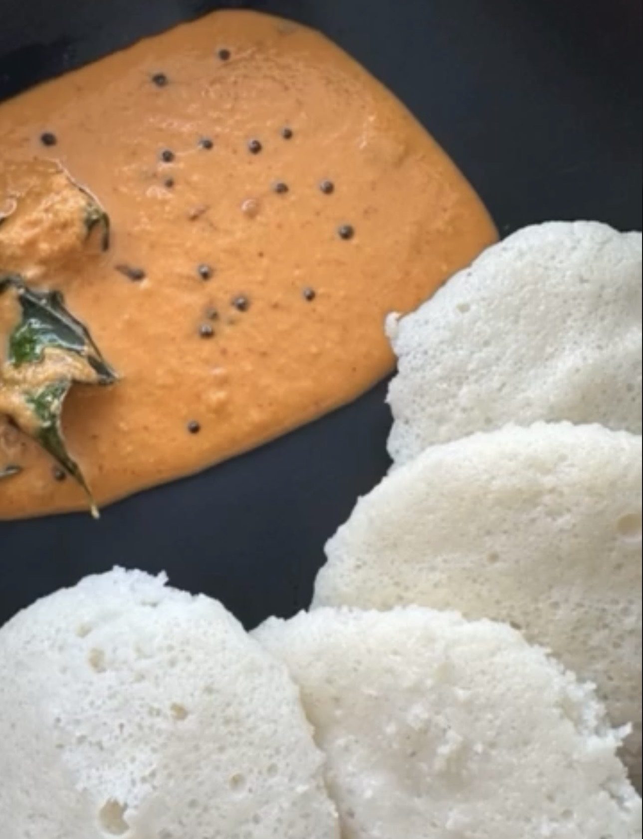 Orange-red Kempu Chutney with mustard seeds and curry leaves beside white idlis on a dark plate
