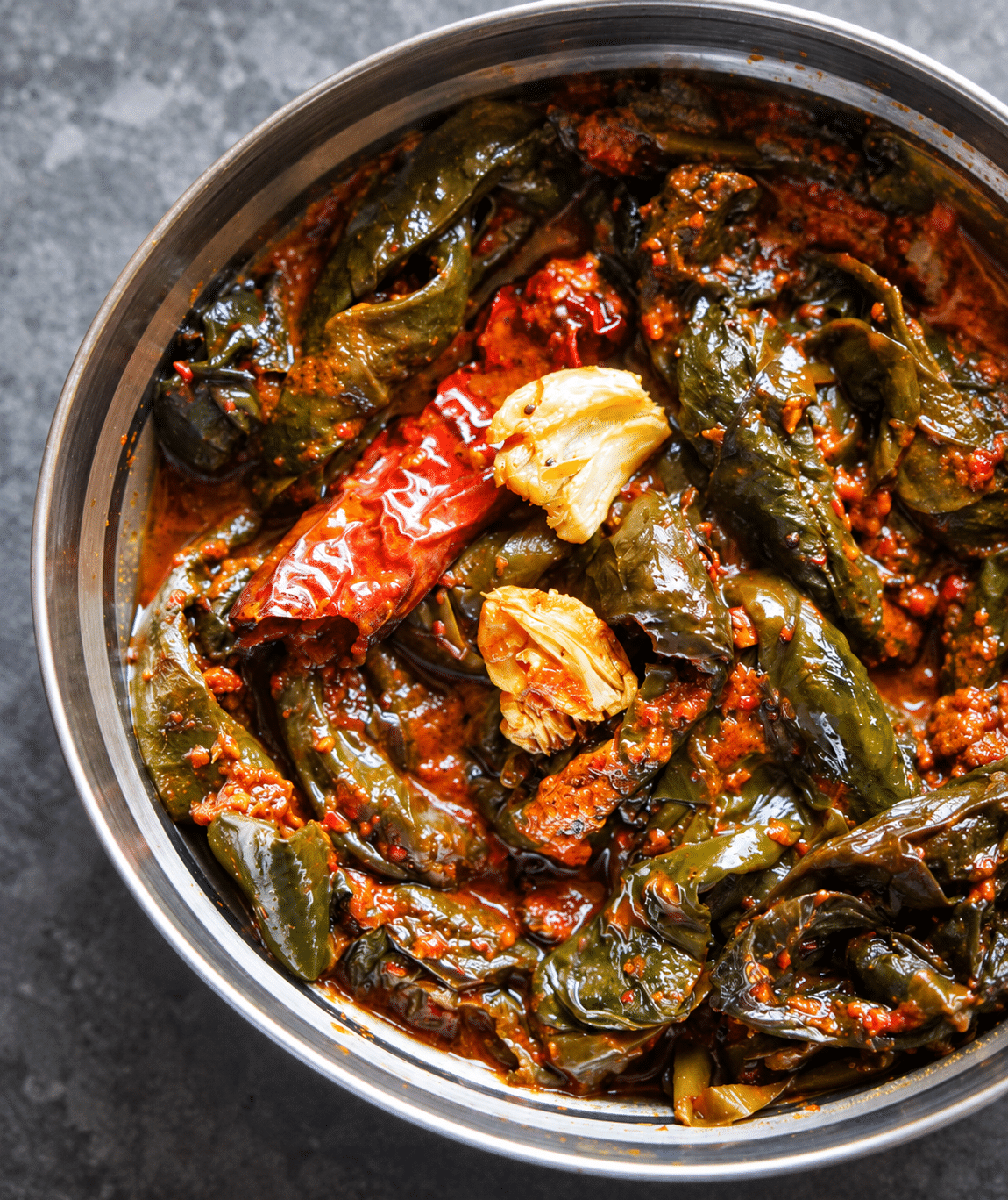 Cooked colocasia leaf knots in red Pulimunchi masala, topped with browned garlic and a dried red chilli in a steel bowl.